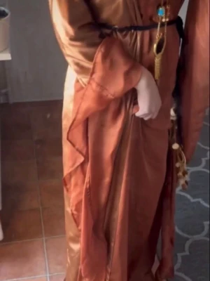 Kurdisk klänning - Kurdisk klänning i skimrande kopparfärgad satin med vida ärmar och guldfärgade detaljer. Klänningen har ett elegant bälte med dekorativa mynt.