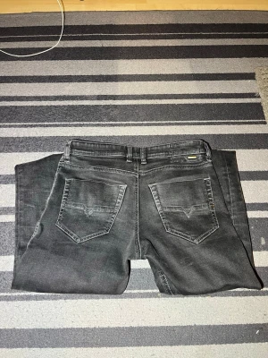 Svarta jeans från Diesel - Säljer ett par svarta jeans från Diesel med klassisk femficksmodell och snygga slitningar. Jeansen har normal passform och är tillverkade i bomull med en liten mängd stretch för extra komfort. Perfekta till en avslappnad streetstyle-look.