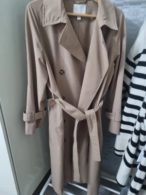 Beige trenchcoat från H&M - Stilren beige trenchcoat från H&M med klassisk dubbelknäppning, bred krage och avtagbart midjebälte. Jackan har långa ärmar med justerbara slejfar vid handlederna och är tillverkad i ett mjukt, vävt material. Perfekt för en trendig och tidlös look.