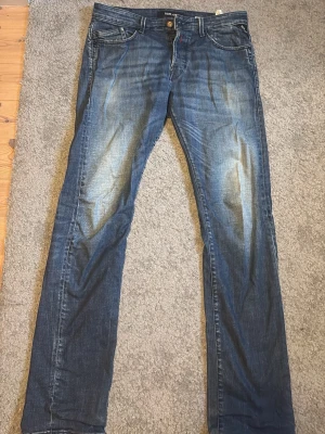 Blå raka jeans från Replay - Säljer ett par blå jeans från Replay med raka ben och klassisk femficksmodell. Jeansen har slitningar och ljusare partier framtill för en cool, använd look. Tillverkade i denim med synliga sömmar och knappgylf.