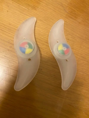 Cykelhjul LED-ljus clips - Två genomskinliga LED-ljus clips för cykelhjul. Clipsen har en böjd form och fästs enkelt mellan ekrarna. I mitten finns en rund del med färgade sektioner som lyser upp när hjulet snurrar. Perfekt för att synas bättre i mörker och ge cykeln en cool look.