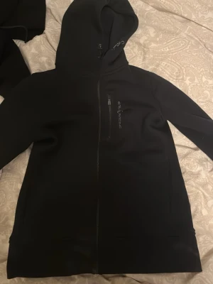 Svart hoodie från Sail Racing - Svart hoodie från Sail Racing med dragkedja framtill och en bröstficka med logga. Hoodien har en klassisk huva med dragsko och är tillverkad i ett mjukt och lite tjockare material. Perfekt för dig som gillar stilren och sportig design.