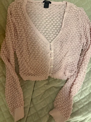 Ljusrosa virkad kofta från Lindex - Supersöt ljusrosa kofta från Lindex med virkat mönster och v-ringning. Koftan har långa ärmar, ribbade muddar och knäpps med fem knappar framtill. Kortare modell som passar perfekt till högmidjade jeans eller kjol eller ny till våren💕