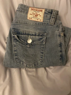 True Religion Becca Bootcut Jeans - Säljer ett par ljusblå jeans från True Religion, modell Becca Mid Rise Bootcut. Jeansen har klassiska kontrastsömmar, ficklock med knapp bak och en snygg bootcut passform. Tillverkade i denim med en cool tvättad look och detaljerade sömmar.
