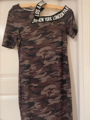 Camouflage klänning med textband - Cool kortklänning i camouflage-mönster med svartvitt textband över ena axeln där det står 'LOS ANGELES NEW YORK LONDON PARIS'. Klänningen har korta ärmar och är figurnära. Perfekt för dig som vill sticka ut med streetstyle-vibe.