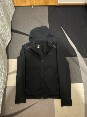 Svart jacka från Under Armour - Snygg svart jacka från Under Armour med dragkedja framtill och huva. Diskret logga på bröstet och stor Under Armour-text på ryggen. Tillverkad i ett mjukt och stretchigt material som känns sportigt och bekvämt. Perfekt för dig som gillar en clean och stilren look.