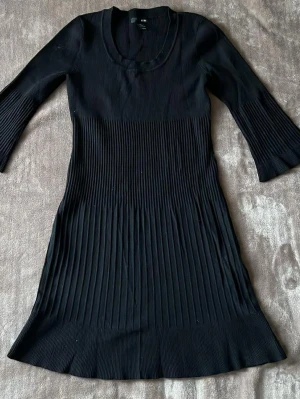 Svart ribbad klänning från H&M - Svart ribbad klänning från H&M med rund halsringning och trekvartsärm. Klänningen har en figurnära passform upptill och lätt utsvängd nederdel. Materialet är stretchigt och bekvämt, perfekt för en stilren look.