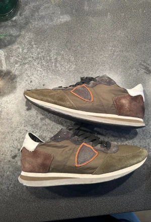 Sneakers från Philippe Model i olivgrönt - Snygga sneakers från Philippe Model i olivgrönt tyg och mocka med bruna och vita detaljer. Skorna har svarta skosnören, vit sula och en orange kant runt logotypen på sidan. Perfekta för dig som gillar en sportig och trendig look.