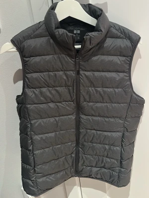 Grå dunväst från Uniqlo - Snygg grå dunväst från Uniqlo med lättviktsdesign och horisontella sömmar. Västen har hög krage och dragkedja framtill, perfekt för lager-på-lager-stil. Tillverkad i ett tunt, smidigt material som håller värmen utan att kännas klumpig.