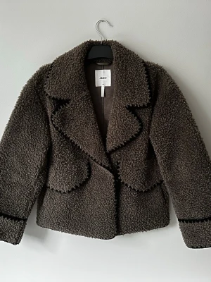 Brun teddyjacka från Object - Mysig brun teddyjacka från Object med bred krage och markerade sömmar. Jackan har en boxig passform och är tillverkad i ett fluffigt teddy-material som ger en cool och trendig look. Perfekt för kyliga dagar när du vill vara både varm och snygg.