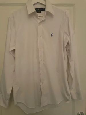 Vit skjorta från Ralph Lauren - Klassisk vit skjorta från Ralph Lauren med blå logga broderad på bröstet. Skjortan har lång ärm, knappar framtill och klassisk krage. Perfekt för en clean och stilren look.
