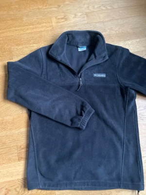 Svart fleece från Columbia - Mjuk och varm svart fleece från Columbia med halv dragkedja framtill och hög krage. Perfekt för kyliga dagar och enkel att matcha med andra plagg. Tillverkad i skönt fleecematerial och har diskret logga på bröstet.