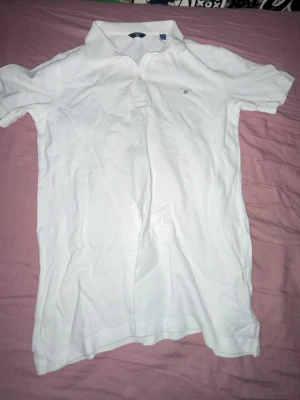 Vit pikétröja från GANT - Klassisk vit pikétröja från GANT med liten broderad logga på bröstet. Tröjan har krage, knappar framtill och är tillverkad i ett mjukt bomullsmaterial. Perfekt för en clean och stilren look.