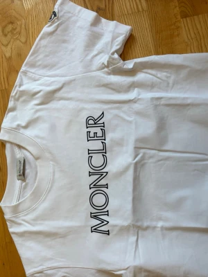 Vit Moncler t-shirt med logga - Vit t-shirt från Moncler med stor svart logotyp tryckt vertikalt på framsidan. Klassisk rund halsringning och korta ärmar. Tillverkad i mjuk bomull för en bekväm känsla och stilren look.