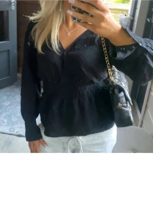 Svart v-ringad blus  - Snygg svart blus med v-ringning och markerad resår i midjan som ger en fin siluett. Blusen har långa ärmar med knapp vid ärmslut och är tillverkad i ett lätt tyg som känns skönt mot huden. Perfekt att matcha med jeans eller kjol.