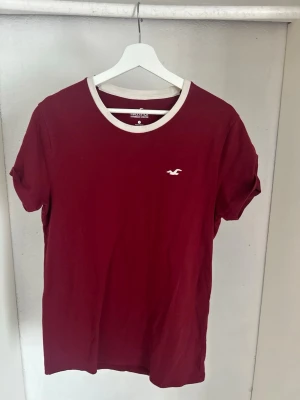 Vinröd t-shirt från Hollister - Snygg vinröd t-shirt från Hollister med vit kant runt halsen och liten logga på bröstet. Klassisk passform och mjukt bomullsmaterial gör den perfekt till alla tillfällen. Enkel sopran look som funkar till det mesta.