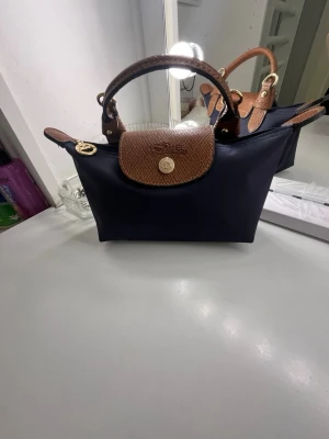 Mörkblå handväska från Longchamp KOPIA - Snygg mörkblå handväska från Longchamp med bruna läderdetaljer på handtag och lock. Väskan har guldfärgade metalldetaljer och klassisk knappstängning framtill. Perfekt storlek för det viktigaste och enkel att bära med sig. Det är en kopia men köpte den för 4000kr säljer för 1000kr