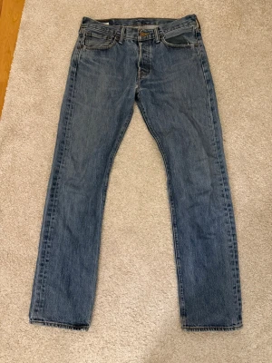 Levis 501 w31/32 - Klassiska blå Levi's jeans med rak passform och fem fickor. Jeansen har en tidlös tvätt och är tillverkade i slitstark denim. Perfekta för en avslappnad och cool stil. Levi's-lapp bak i midjan och ikonisk röd etikett på bakfickan.