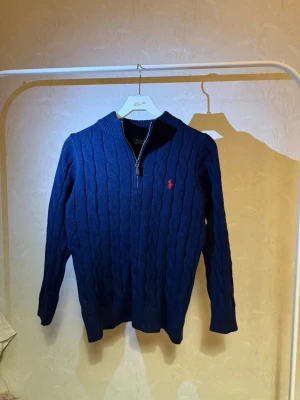 Marinblå stickad tröja Polo Ralph Lauren - Mörkblå stickad tröja från Polo Ralph Lauren med halv dragkedja och det klassiska röda logobroderiet på bröstet. Tröjan har kabelstickat mönster, ribbade muddar och hög krage. Perfekt för lager på lager och snygg till jeans.