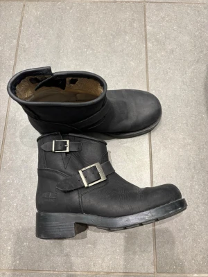 Svarta biker boots med spännen Johnny Bulls - Snygga svarta biker boots i läder med två silverfärgade spännen och rund tå. Grov sula och låg klack ger en cool look. Insidan är fodrad för extra komfort. Perfekta till jeans eller skinnjacka.