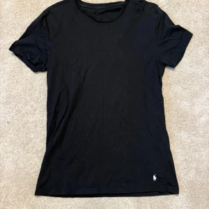 Svart t-shirt från Polo Ralph Lauren - En klassisk svart t-shirt från Polo Ralph Lauren med korta ärmar och rund hals. Diskret broderad logga i vitt på nedre delen av framsidan. Perfekt för en clean och enkel look, tillverkad i mjuk bomull.