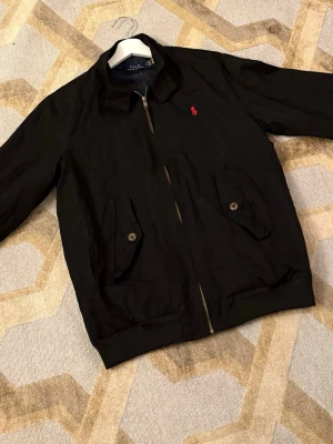 Svart jacka från Polo Ralph Lauren - Klassisk svart jacka från Polo Ralph Lauren med dragkedja, två fickor med lock och knappar samt röd broderad logga på bröstet. Jackan har en enkel krage och ribbade muddar vid midjan. Perfekt för dig som gillar stilren och tidlös design.