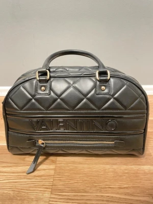 Svart quiltad handväska Valentino - Snygg svart handväska från Mario Valentino med quiltat mönster och guldfärgade  detaljer. Väskan har ett stort VALENTINO-tryck framtill, dragkedja och dubbla handtag. Aldrig använd, fick den som 18års present, inte riktigt min stil. 