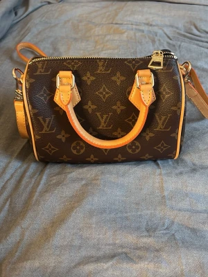 Louis Vuitton Speedy Bandoulière 20 - Säljer en brun Louis Vuitton Speedy Bandoulière 20 med klassiskt monogrammönster i canvas och detaljer i beige och orange läder. Väskan har guldfärgade metalldetaljer, dubbla handtag och avtagbar axelrem. Perfekt för dig som gillar lyxiga accessoarer. Är villig att diskutera pris