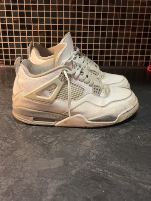 Vita oreo Jordan 4 - Säljer ett par klassiska Air Jordan 4 sneakers i vitt med grå detaljer och röd Jumpman-logga på plösen. Skorna har meshpaneler, plastdetaljer och synlig Air-dämpning i sulan. Perfekt för dig som gillar retrostil och ikoniska sneakers.