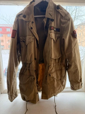 Beige fältjacka från Polo Ralph Lauren - Säljer en beige fältjacka från Polo Ralph Lauren med flera fickor, militärinspirerade patchar och snörning i midjan. Jackan har krage, axelklaffar och dragkedja samt knappar framtill. Perfekt för dig som gillar utility-stil och detaljer.