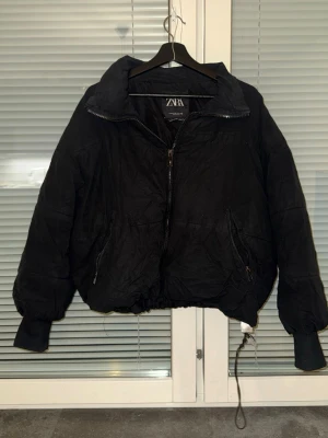 Svart pufferjacka från Zara - Svart pufferjacka från Zara med hög krage, dragkedja framtill och två snedställda fickor med dragkedja. Jackan har resår i midjan och vid ärmsluten samt justerbar dragsko nedtill. Perfekt för kalla vinterdagar.
