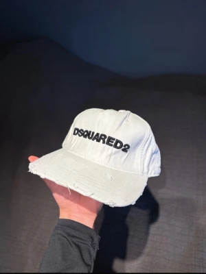 Vit Dsquared2 keps med brodyr - Snygg vit keps från Dsquared2 med svart broderad logga framtill och texten 'Dean & Dan Caten' baktill. Kepsen har böjd skärm, justerbart spänne och slitna detaljer för en trendig look. Perfekt accessoar för att lyfta din outfit.