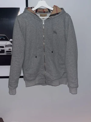 Burberry Zip - Snygg grå hoodie med dragkedja från Burberry. Klassisk huva med det ikoniska rutmönstret på insidan, dragsnören och två fickor framtill. Perfekt för en chill och stilren look. Materialet är mjukt och bekvämt, passar till det mesta. Storleken är S och den sitter bra på mig som är 169cm lång! Väldigt fint skick😁