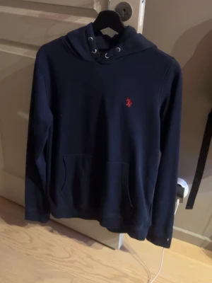Mörkblå hoodie från Ralph Lauren - Mörkblå hoodie från Ralph Lauren med klassisk huva och känguruficka framtill. Liten röd broderad logga på bröstet. Mjuk bomullskänsla och stilren design som passar perfekt till jeans eller joggers. Typ aldrig använd