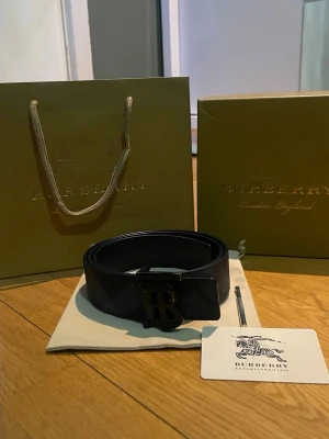 Svart Burberry bälte med monogramspänne - Snyggt svart bälte från Burberry med klassiskt rutmönster och ett stort monogramspänne i metall. Bältet är gjort i läder och har en exklusiv känsla, perfekt för att lyfta vilken outfit som helst. Kommer med originalpåse och Burberry-förpackning. 95 cm