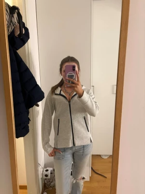 Grå zip-hoodie med svarta detaljer - Säljer en ljusgrå hoodie med dragkedja och svarta detaljer vid fickor och zip. Hoodien har långa ärmar, två fickor framtill och en huva. Perfekt för chill dagar och enkel att matcha med jeans eller joggers.