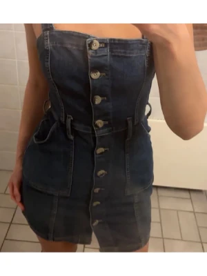 Jeansklänning - Snygg mörkblå jeansklänning med knappar hela vägen framtill och tillhörande bälte i midjan. Klänningen är ärmlös med breda axelband och har praktiska fickor på sidorna. Perfekt för dig som gillar en cool och trendig denimlook.