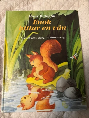 Enok hittar en vän - En charmig barnbok med hårda pärmar, skriven av Hans Wilhelm och svensk text av Birgitta Rosenberg. Omslaget har en färgglad illustration av en ekorre vid en damm, tillsammans med en groda och en trollslända. Boken har ett lekfullt och varmt uttryck.