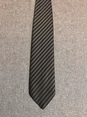 Randig slips från Jens Engwall - Snygg slips i svart och grått med diagonala ränder från Jens Engwall. Tillverkad i ett glansigt tyg som ger en elegant känsla. Perfekt accessoar för att lyfta din outfit.