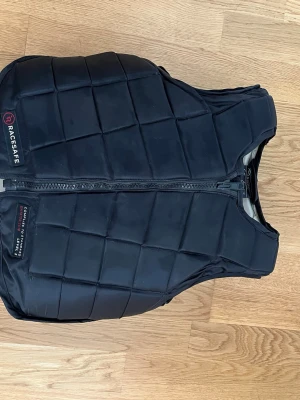 Svart skyddsväst från Racesafe - Säljer en svart skyddsväst från Racesafe med rutmönstrad vaddering och dragkedja framtill. Västen har justerbara band och är sparsamt använd. Har aldrig ramlat av i den. Nypris är ca 3000kr då det är den senaste modellen. Storlek large för barn vilket även fungerar som xs för vuxen.