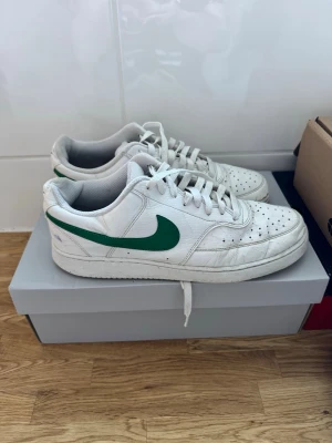 Nike vita sneakers med grön swoosh - Fräscha vita Nike sneakers med klassisk grön swoosh på sidan. Lågt skaft, perforerad tåbox och vit yttersula med Nike-logga undertill. Skorna har en stilren och sportig look, perfekta till streetwear. Ovandelen är i läder och de har vita skosnören.