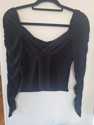 Svart omlottopp från Bik Bok - Snygg svart omlottopp från Bik Bok med långa, lätt puffade ärmar och djup v-ringning. Toppen har en croppad passform och är tillverkad i ett mjukt, stretchigt material som sitter bekvämt. Perfekt för dig som vill ha en trendig och stilren look.