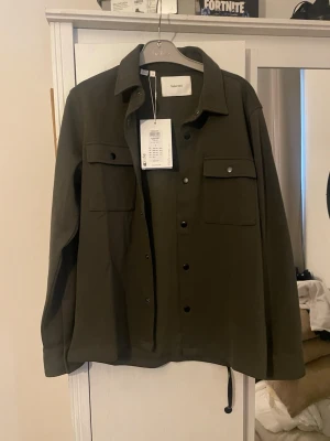 Mörkgrön overshirt från Selected - Stilren mörkgrön overshirt från Selected med klassisk krage och tryckknappar framtill. Två bröstfickor med lock och knappdetaljer ger en snygg look. Tillverkad i ett mjukt tyg som passar perfekt till lager-på-lager-stil.