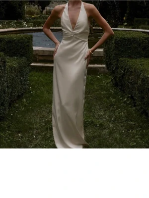 Champagnefärgad långklänning i satin - Elegant långklänning i champagnefärgad satin med djup v-ringning, öppen rygg och slits bak. Klänningen har smala axelband som knyts i nacken och en figurnära passform som ger en lyxig känsla.