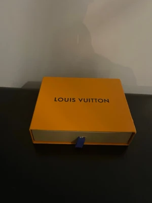  svart monogram plånbok - Snygg svart plånbok från  med klassiskt monogrammönster i grått. Tillverkad i exklusivt läder och levereras i original orange box med blått band. Perfekt för dig som vill ha en stilren och lyxig accessoar.