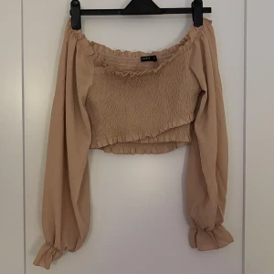Beige offshoulder croppad topp Gina Tricot - Trendig beige offshoulder topp från Gina Tricot med croppad passform. Toppen har långa puffiga ärmar med resår vid ärmslut och smockad kropp för en snygg och bekväm look. Perfekt att styla med höga jeans eller kjol.