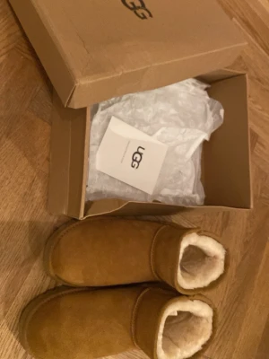 Bruna fårskinnsskor från UGG - Mysiga bruna skor från UGG i klassisk modell med mjukt fårskinnsfoder och ovandel i mocka. Perfekta för att hålla fötterna varma. Skorna skickas i orginallåda.