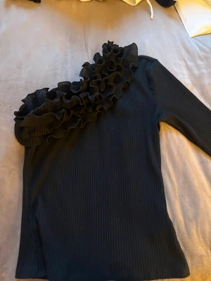 Svart oneshoulder topp med volang - Snygg svart oneshoulder topp med ribbad struktur och stora volangdetaljer längs ena axeln. Toppen har lång ärm på ena sidan och ärmlös på andra, vilket ger en unik och trendig look. Perfekt för dig som vill sticka ut lite extra.
