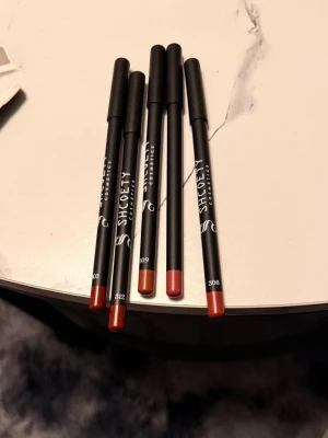 Shoety Cosmetics läppennor mix - Fem snygga läppennor från Shoety Cosmetics i olika nyanser av rött, brunt och nude. Pennorna har svart hylsa med färgkodad ände och tydlig logga. Perfekta för att definiera och forma läpparna med precision.