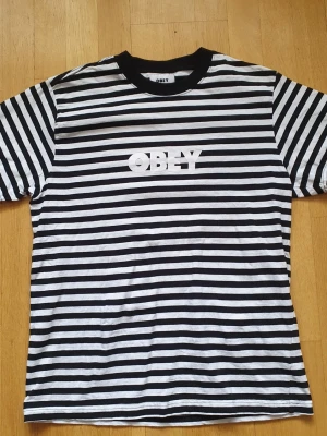 Obey T-Shirt | M / Medium - T-Shirt från Obey med tryck på framsidan.  Medium (Mått finns på bilderna!) 100% bomull.  Se bilder.  "Obey Clothing är ett amerikanskt streetwear-företag grundat 2001 av gatukonstnären och illustratören Shepard Fairey som en förlängning av hans arbete inom street art och aktivism. Bilderna på märkeskläderna varierar brett och inkluderar motiv som parodierar massmarknadskonsumtion"